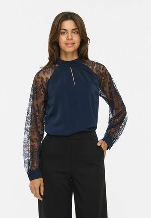 Une femme porte un chemisier bleu marine à manches en dentelle transparente et un pantalon noir sur un fond blanc uni.