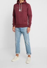 Sudadera burdeos con un bolsillo frontal y cordones; presenta el logo y emblema de "GANT". Combinada con unos vaqueros de puño enrollado azul claro y zapatillas blancas.