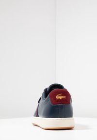 Granatowy skórzany sneaker z bordowym zamszowym obcasem, białą podeszwą i złotym logo. Posiada niską cholewkę oraz kontrastowe paski.