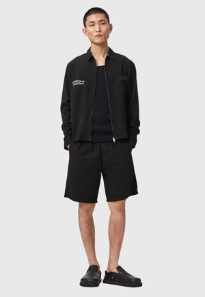 Ung mand stående iført en sort lynlåstrøje med hvid tekst, sorte shorts og sorte slip-on sko, foran en ensfarvet lys grå baggrund.