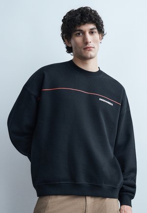 Fiatal férfi göndör hajjal, fekete DSQUARED2 pulóvert visel piros vonalrészlettel és bézs nadrágot, világosszürke háttér előtt áll.