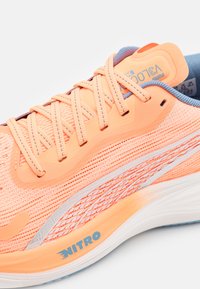 Zapatilla de running Puma con una parte superior de malla naranja transpirable, textura intrincada, detalles en blanco y la marca 'NITRO' en azul en la suela.