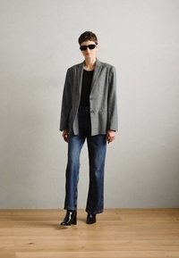 Gray blazer, black top, blue wide-leg jeans, and black ankle boots. Model wearing black sunglasses against a light background.