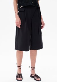 Zwarte wijde shorts met een gladde textuur, voorzien van een strik in de taille, gecombineerd met zwarte geweven sandalen en een opengehaakt topje.