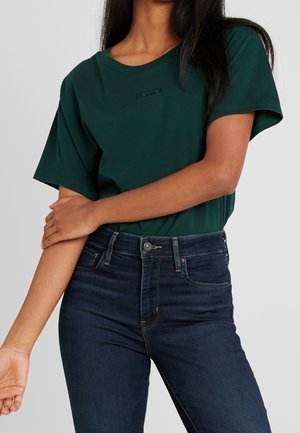 Une personne avec de longs cheveux foncés porte un T-shirt vert foncé et un jean en denim sombre, les bras croisés dans un cadre neutre.