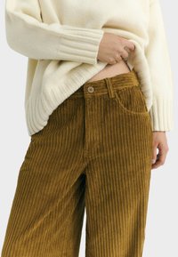 Person lifting cream sweater, revealing midriff, wearing high-waisted mustard corduroy pants with button and front pockets.