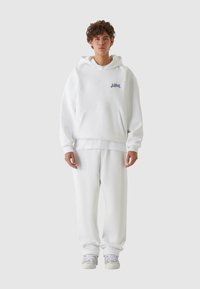 Hvit hettegenser med frontlomme og liten blå logo, kombinert med matchende hvite joggebukser og lys grå joggesko, som viser en avslappet passform.