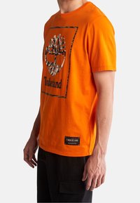 Hombre vistiendo una camiseta de Timberland naranja brillante con el logo del árbol de camuflaje y pantalones cargo negros, mostrado de lado, contra un fondo liso.