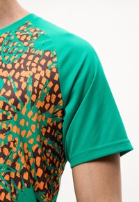 Πράσινο πουκάμισο με κοντά μανίκια και πορτοκαλί και μαύρο λουλουδάτο σχέδιο στο πάνω μέρος του στήθους και στις ώμοι.