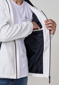 Jachetă albă cu interior negru, dotată cu un fermoar și o buzunar interior. Materialul exterior are o textură netedă, concepută pentru confort.