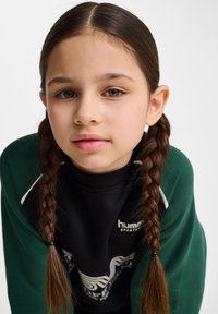 Grön tröja över en svart topp med vita accenter, med flätor i håret, neutral bakgrund och fokuserat ansiktsuttryck.