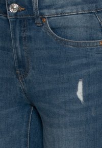 Blue denim jeans showing front pocket, button closure, belt loop, and small vertical distress detail near the thigh.