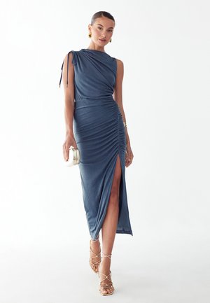 Vrouw in een mouwloze, geplooide blauwe midi-jurk met een split aan de zijkant, gouden oorbellen, sandalen met bandjes en een metallic clutchtasje in de hand.