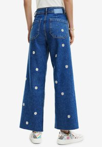 Wide-leg jeans in blue denim, featuring white daisy embroidery. Two back pockets and a branded waistband accentuate the design.