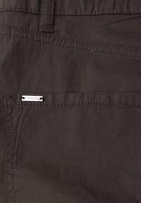 Donkerbruine stoffen zak met zichtbare stiksels en een klein rechthoekig metalen merklabel bevestigd vlakbij de bovenrand van de zak.