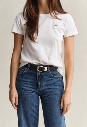 Kvinde iført en simpel hvid t-shirt med korte ærmer, stoppet ned i højttaljede blå jeans med sort bælte og guldfarvet spænde.
