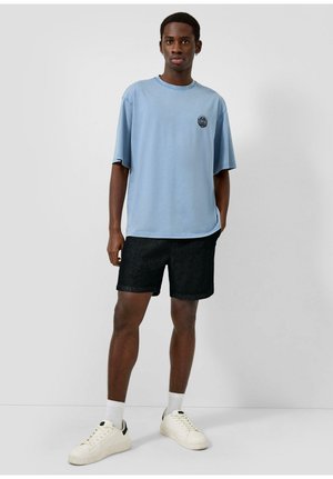 Młody mężczyzna stoi przy gładkiej ścianie, ubrany w jasnoniebieski oversize T-shirt, czarne spodenki, białe skarpetki i białe tenisówki.