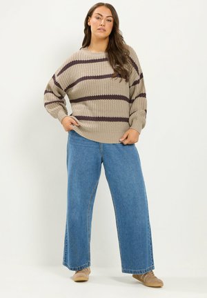 Femme debout les mains dans les poches portant un pull rayé beige et marron, un jean large bleu et des baskets beige clair sur fond blanc.