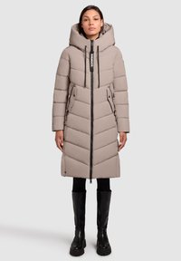 Vrouw in een knielange beige pufferjas met een grote capuchon, rits aan de voorkant, klepzakken, zwarte leggings en zwarte kniehoge laarzen.