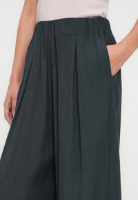 Weit geschnittene Hose aus dunkelgrünem, strukturiertem Stoff. Verfügt über einen gerafften Taillenbund und Seitentaschen. Falten verleihen Form und Dimension.