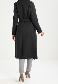 Cappotto lungo grigio scuro con cintura, caratterizzato da un colletto a revers e taglio dritto. Indossato sopra pantaloni grigio chiaro e tacchi grigi. Tessuto dalla trama liscia.
