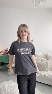 Νεαρή γυναίκα με ξανθά μαλλιά που φοράει σκούρο μπλουζάκι με το λογότυπο "Rip Curl Australia" και μαύρο παντελόνι, στέκεται σε ένα φωτεινό σαλόνι.