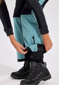 Water-resistant hiking pants in teal and black, featuring adjustable cuffs and zippered pockets, worn with black hiking boots.
