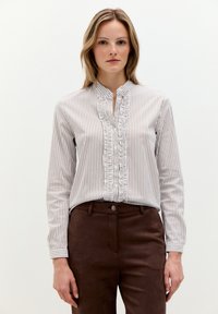 Weißes Bluse mit beigen vertikalen Streifen, ausgestattet mit einer rüschigen Frontleiste und langen Ärmeln. Kombiniert mit braunen Hosen. Glatte Textur.