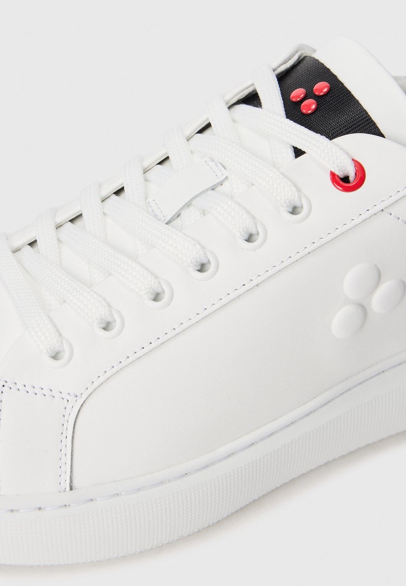 Sneaker bianco con lacci bianchi, lingua nera, tre punti rossi vicino all'occhiello superiore e tre punti bianchi in rilievo sul pannello laterale.