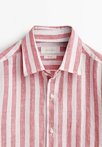 Chemise en lin à rayures verticales rouges et blanches, dotée d'un col pointu, d'une fermeture à boutons sur le devant, et d'une étiquette indiquant un matériau 100% lin.