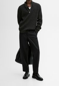 Homme portant un pull côtelé foncé, un pantalon large noir, des chaussures en cuir noires, tenant un sac noir sur un fond blanc uni.