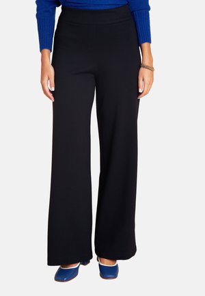 Frau trägt hochgeschnittene schwarze weit geschnittene Hosen, blaues langärmeliges Oberteil, blaue flache Schuhe mit quadratischer Spitze, mit goldenen Armbändern am rechten Handgelenk.