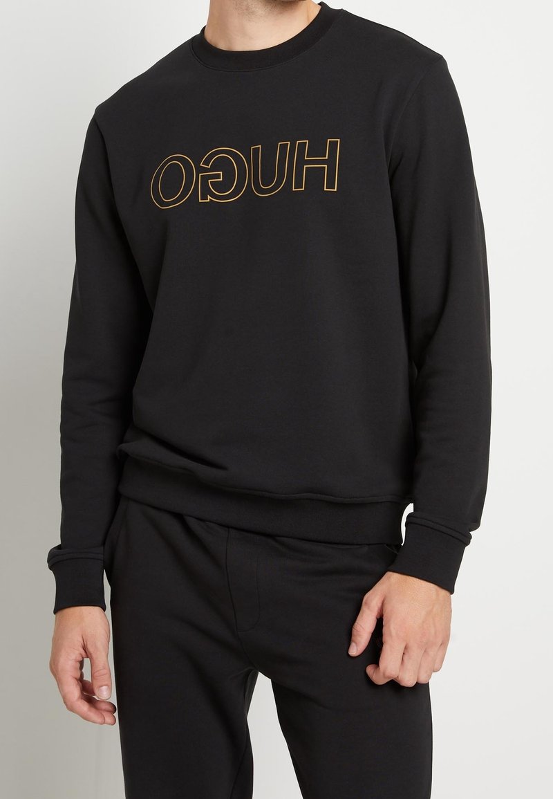 Homme portant un sweat-shirt noir avec un texte doré inversé "HUGO" et un pantalon noir assorti, debout devant un fond clair.