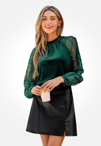 Groene fluwelen blouse met kanten mouwen, geplooide halslijn, gecombineerd met een zwarte nepleer rok met een zijsplit. Houdt een kopje vast.