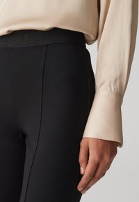 Pantalons noirs ajustés avec une taille texturée, associés à un chemisier long à manches longues beige doux avec des poignets boutonnés.
