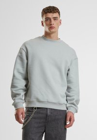 Hellgrauer Sweatshirt aus weichem Material, mit rundem Ausschnitt und gerippten Bündchen. Kombiniert mit dunkelgrauen Jeans mit einem Metalldetail in Form einer Kette.
