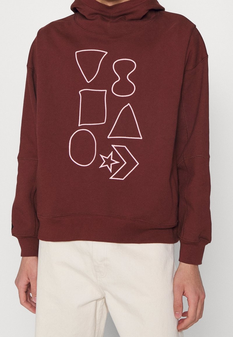 Personne portant un sweat à capuche bordeaux avec des formes géométriques blanches en contour, comprenant un triangle, un cercle, un carré, une étoile et une flèche imprimées sur le devant.