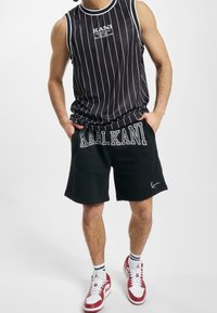 Canotta nera a righe con logo, abbinata a pantaloni corti neri con testo stampato bianco. Sneakers bianche e rosse completano l'outfit.