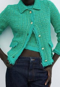 Turkusowy sweter kardigan o fakturowanej powierzchni z postrzępionymi krawędziami, zapinany na guziki, z dopasowanym dzianinowym topem. Do tego ciemne dżinsy.