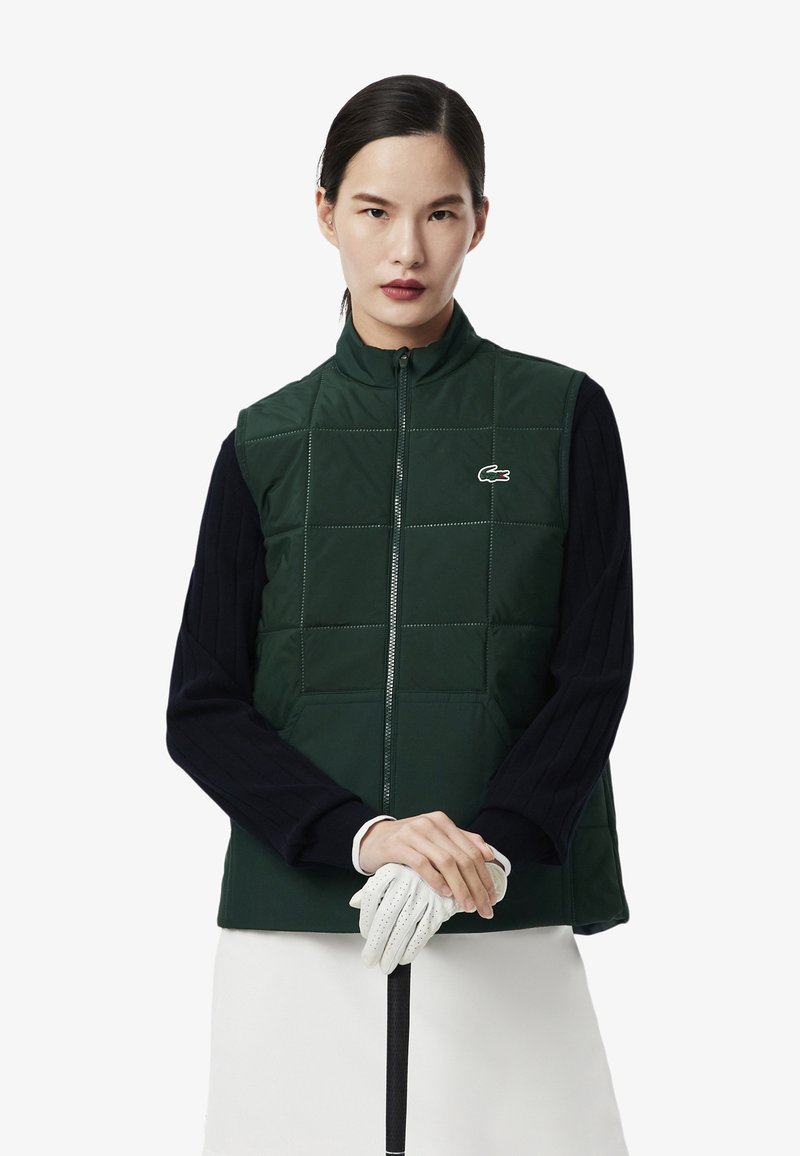 Donkergroene puffer vest met een doorgestikte ontwerp, hoge kraag en rits aan de voorkant; gedragen over een marineblauw longsleeve shirt, met een golfclub en handschoen vast.