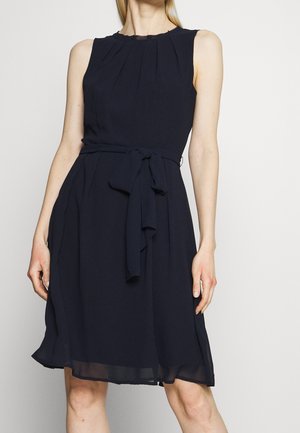 Czarna, bezrękawna sukienka do kolan z wiązanym paskiem w talii i plisowanym dekoltem, noszona przez stojącą osobę na jednolitym tle.