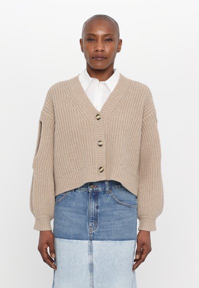 Beige gestrickte Strickjacke mit V-Ausschnitt, kürzerer Länge, mit vier Knöpfen und geripptem Muster. Über einem Jeansrock getragen.