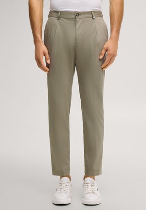 Mann trägt beige gerade geschnittene Hosen mit vorderen Falten, Knopf- und Reißverschluss sowie weißen Turnschuhen vor neutralem Hintergrund.