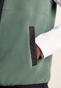 Fleece jacket in green with a textured surface, featuring a black pocket trim and a white sleeve peeking from behind.