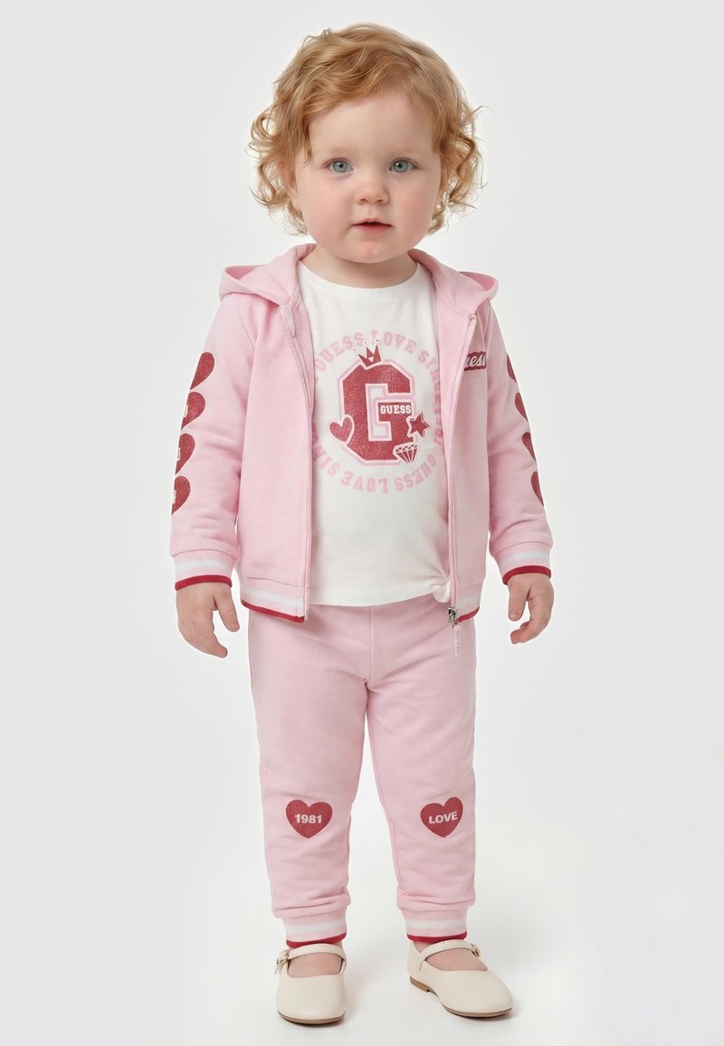 Tout-petit aux cheveux roux bouclés portant une veste à capuche rose et un pantalon avec des motifs de cœurs rouges, une chemise blanche et des chaussures crème, debout sur fond blanc.