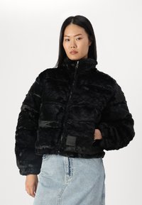 Jachetă scurtă din blană artificială neagră, cu guler înalt, închidere cu fermoar și design matlasat. Textură moale și croială lejeră, având un patch cu logo.