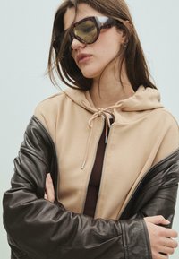 Žena v overzise slunečních brýlích ve želvovinovém stylu, béžové mikině s šňůrkami a částečně rozepnuté černé kožené bundě, se založenýma rukama.
