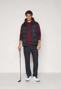 Ανδρικό σύνολο με μαύρο φουσκωτό γιλέκο πάνω από μπορντό φούτερ, σκούρους παντελόνες και λευκά γκολφ παπούτσια με μπλε λεπτομέρειες, κρατώντας ένα μπαστούνι του γκολφ.