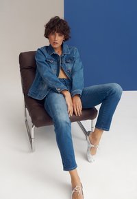 Denim jacket and jeans in medium blue, worn with silver flats. Sitting on a brown chair, minimal background with blue and beige hues.