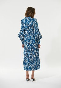 Blaues Blumenkleid mit gestuften Saum, langen Puffärmel und tailliertem Schnitt. Hergestellt aus einem glatten Stoff mit einem lebendigen weißen und blauen Muster.